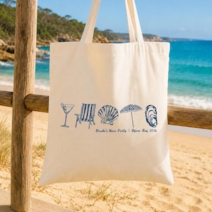 May include: A cream-coloured canvas tote bag featuring a blue graphic print of a martini glass, a beach chair, a seashell, a beach umbrella, and an oyster shell, with the text "Brooke's Hens Party | Byron Bay 2026" printed below the icons.