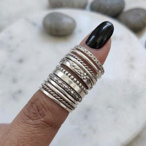 May include: A stack of nine silver rings worn on a finger, featuring various textures including twisted rope patterns, smooth polished bands, and intricate floral or geometric engravings, creating a layered metallic appearance on the finger.