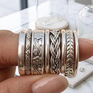 May include: A set of seven silver-toned rings stacked together. The collection includes plain bands, a band with a geometric diamond pattern, a floral embossed band, a braided rope-style band, and a band featuring a repeating leaf-like pattern.