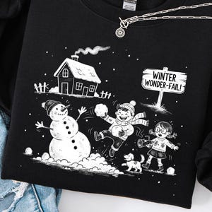 Könnte beinhalten: Schwarzes Sweatshirt mit einem winterlichen Grafikdesign. Die Grafik zeigt einen Schneemann, Kinder, die im Schnee spielen, ein Haus und den Text "Winter Wonder-Fail!". Eine silberne Halskette liegt über dem Sweatshirt.