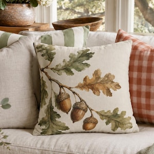 Op de afbeelding: Een vierkant sierkussen met een botanische illustratie van bruine eikels en groene en bruine eikenbladeren op een crèmekleurige achtergrond, geplaatst op een bank naast een effen beige kussen en een oranje-wit geblokt kussen.