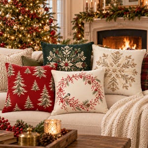 May include: A collection of square throw pillows featuring holiday designs. One is red with green pine trees, one is dark green with a white snowflake, one is cream with a gold snowflake, and one is cream with a red berry wreath and a bird.