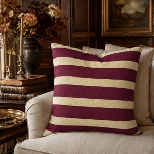May include: A square scatter cushion featuring a repeating horizontal striped pattern in deep burgundy and cream. The fabric has a visible woven texture, and the cushion is positioned upright on a neutral-coloured upholstered chair.