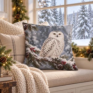 May include: A square throw pillow featuring a detailed print of a white snowy owl perched on a pine branch covered in snow and red berries. The background depicts a soft-focus winter forest scene with falling snow, set against a neutral fabric.