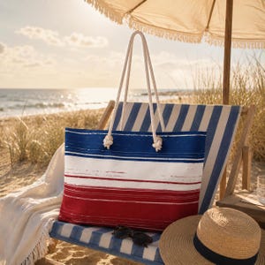 Peut inclure: Un sac fourre-tout en toile avec un motif à rayures bleues, blanches et rouges, avec des poignées en corde. Le sac est sur une chaise longue à rayures bleues et blanches, avec un chapeau de paille et des lunettes de soleil à proximité.