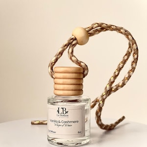 May include: A small, clear glass car air freshener with a wooden cap and a braided cord. The label reads "Vanilla & Cashmere Whisper of Winter" and "Car Diffuser 8ml". The cord is tan and gold. The wooden cap is made of stacked rings.