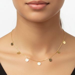 Könnte beinhalten: Eine goldfarbene Halskette mit mehreren kleinen, runden Anhängern. Einige Anhänger haben einzelne Buchstaben. Die Halskette wird um den Hals getragen und liegt auf der Brust. Der Hintergrund ist ein schwarzes Oberteil.