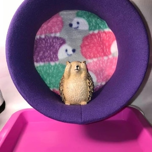 May include: A purple, circular pet bed with a patterned interior featuring smiling hedgehog faces in pink, green, and purple. A small hedgehog figurine is inside the bed. A bright pink tray is in the foreground.
