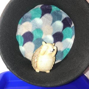 Puede incluir: Una cama para mascotas de fieltro negro con un forro polar estampado en tonos turquesa, azul y gris. Una pequeña figura de erizo marrón claro se coloca sobre el forro polar. La cama está dentro de un recipiente de plástico azul.