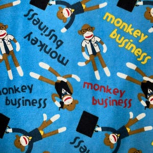 Puede incluir: Tela azul con un patrón repetido de ilustraciones de monos calcetín con atuendo de negocios, sosteniendo maletines. Las palabras "monkey business" están dispersas por el diseño en rojo y amarillo.