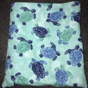 Puede incluir: Una manta azul claro con un estampado de tortugas marinas en tonos azules y verdes. Las tortugas tienen diseños detallados en sus caparazones y están dispersas sobre la tela suave. La manta probablemente está hecha de un material afelpado.