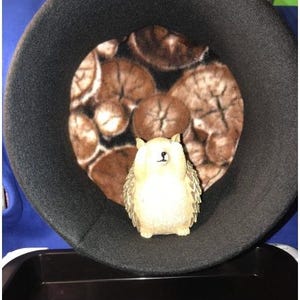 以下が含まれることがあります： 茶色と白の木のスライス模様のインテリアを備えた黒いペットベッド。小さなクリーム色のフクロウの置物がベッドの中にあります。ベッドは丸く、柔らかく豪華な外観をしています。