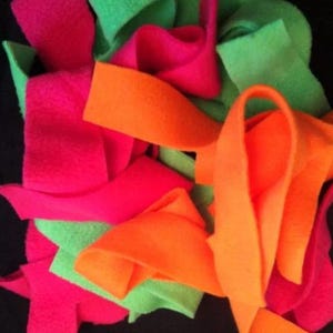 May include: A pile of colourful fleece strips in hot pink, bright green, and orange. The strips are arranged in a haphazard pile, creating a playful and textured appearance. The background is a solid black.