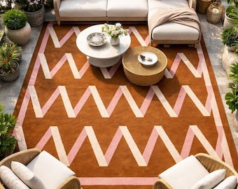 May include: Outdoor patio setting with a large brown rug featuring a zigzag pattern in pink and cream. The scene includes a white sectional sofa with pink pillows, two wicker chairs, and two round coffee tables.