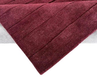May include: A section of a burgundy rug with a textured, ribbed design. The rug is folded over, revealing a lighter gray backing. The angled composition highlights the rich color and plush texture of the rug.