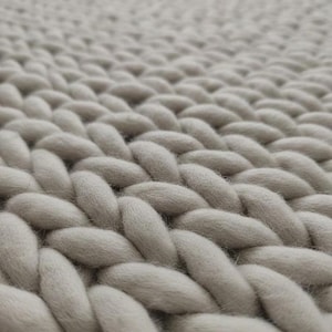 May include: A light gray chunky knit blanket featuring a thick, braided yarn pattern. The texture is composed of large, soft, interlocking loops of yarn that create a consistent, repeating weave across the entire surface of the textile.