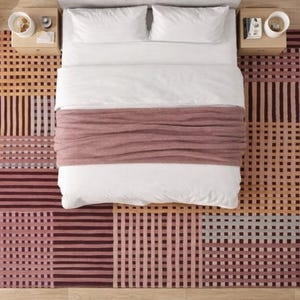 Op de afbeelding: Luchtfoto van een bed met wit beddengoed, een mauvekleurige sprei en een patroonkleed. Het kleed heeft een geometrisch ontwerp in tinten mauve, beige en bruin. Twee nachtkastjes met lampen flankeren het bed. Een potplant en een poef zijn ook zichtbaar.