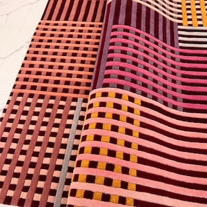 Pode incluir: Tapete enrolado com padrão geométrico. Apresenta uma combinação de listras e quadrados em tons de rosa, bordô e amarelo. O tapete é feito de um material macio e felpudo, ideal para a casa.