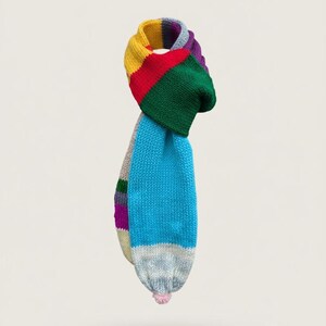 Könnte beinhalten: Ein bunter Strickschal mit Regenbogenmuster. Der Schal hat gelbe, rote, grüne, blaue und violette Abschnitte. Am Ende befindet sich ein hellrosa Bommel. Ein gemütliches Accessoire für kalte Tage.