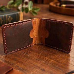 Op de afbeelding: Een bifold portemonnee gemaakt van donkerbruin leer met een verweerde afwerking. De binnenzijde heeft twee kaartsleuven met gebogen uitsparingen en zichtbare stiksels langs de randen, met een lichtere bruine tint in het midden.