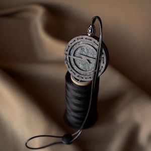 May include: A dark wooden car air freshener with a circular design and a lightning bolt symbol. The air freshener hangs from a black cord and is placed on top of a black spool of thread. The background is a neutral brown.