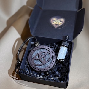 Pode incluir: Um pingente de madeira escura com um design circular e uma inserção de feltro cinza, apresentado em uma caixa de presente preta. A caixa também contém um pequeno frasco com conta-gotas e o texto "À la HU". A caixa tem um adesivo que diz "2026".