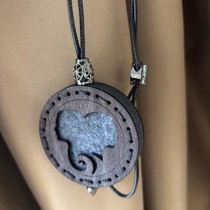 May include: A circular, dark brown pendant necklace with a silhouette design. The pendant has a cutout profile and a grey felt insert. The necklace features a black cord with silver-coloured metal accents. The pendant is approx. 5 cm in diameter.