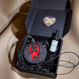 Könnte beinhalten: Eine schwarze Schachtel enthält einen Holzanhänger mit rotem Krebsdesign, eine kleine Flasche und eine schwarze Kordelkette. Der runde Anhänger hat einen detaillierten Krebsausschnitt. Die Flasche hat eine Pipette und ein weißes Etikett. Ein Aufkleber mit dem Text "2026" befindet sich auf der Schachtel.