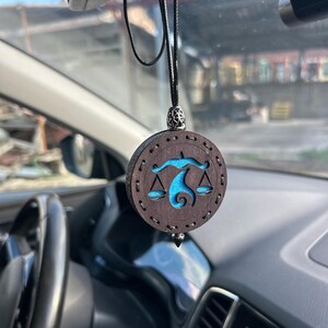 Könnte beinhalten: Ein runder Autoanhänger aus Holz mit dunkelbraunem Finish und einem türkisfarbenen Waage-Symbol. Der Anhänger wird von einer schwarzen Schnur gehalten und hat eine dekorative Metallperle oben und eine silberfarbene Spitze unten.
