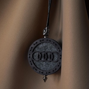 May include: A dark wooden pendant with the Audi logo, featuring a circular design with a grey felt centre. The pendant hangs from a black cord and has a silver-coloured metal accent at the top and a pointed bottom.