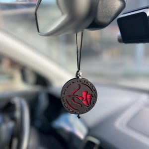 Op de afbeelding: Een zwarte auto luchtverfrisser met een rood Boogschutter ontwerp. De ronde, houten hanger hangt aan een zwart touwtje en heeft een uitgesneden ontwerp van een boogschutter met pijl en boog. De luchtverfrisser hangt aan de achteruitkijkspiegel van een auto.