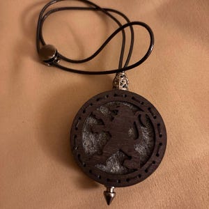 Puede incluir: Un collar con colgante redondo de color marrón oscuro con un diseño de silueta de león. El colgante es de madera y tiene un respaldo de fieltro gris. El collar tiene un cordón negro con un cierre magnético.