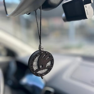 Pode incluir: Um ambientador de carro redondo e preto com o logotipo da Skoda, pendurado por um cordão preto. O ambientador tem um centro de feltro cinza escuro e um topo decorativo prateado. O fundo mostra o interior de um carro, incluindo um espelho retrovisor e uma dashcam.