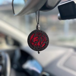 Op de afbeelding: Een ronde auto luchtverfrisser met een leeuwenontwerp. De luchtverfrisser heeft een zwart houten frame met een rode kern en een zilverkleurige bovenkant. Hij hangt aan een zwart touwtje.