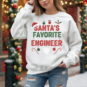 May include: White sweatshirt with the text "SANTA'S FAVORITE ENGINEER" in red and green. The design includes a Santa hat, holly, candy canes, ornaments, and a beard. The sweatshirt is a festive holiday item.