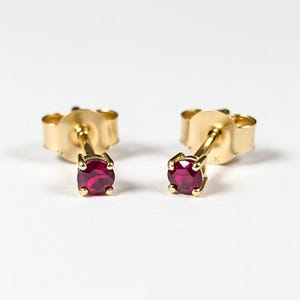 Puede incluir: Un par de pendientes de oro con un diseño clásico, cada uno con una gema redonda de color rojo intenso. Los pendientes tienen un diseño clásico con un poste y un cierre. El oro tiene un acabado pulido.