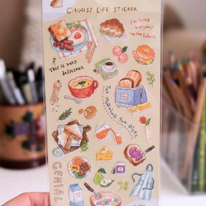 Peut inclure: Une feuille d'autocollants transparente avec des illustrations colorées de plats de petit-déjeuner, notamment des toasts, des œufs et des céréales. Le texte comprend "CHOOSY LIFE STICKER", "This is very delicious", "I'm living everyday to the fullest", "The morning sun rises", "Genial", et "Have you finished your breakfast yet?"