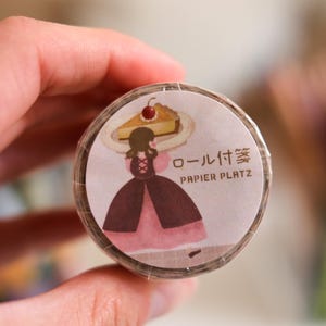 Peut inclure: Rouleau de ruban adhésif décoratif avec une illustration de dessin animé d'une femme portant une part de gâteau. Le ruban adhésif porte le texte "PAPIER PLATZ" et des caractères japonais. Le ruban est marron et rose.