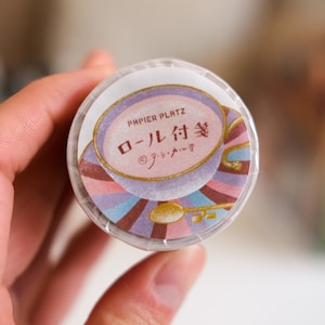 Peut inclure: Un rouleau de ruban adhésif décoratif avec un design coloré. L'étiquette du ruban indique "PAPIER PLATZ" et comporte du texte japonais et une illustration de clé dorée. Le rouleau de ruban adhésif est tenu dans une main.