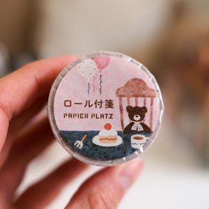 Peut inclure: Un rouleau de ruban adhésif décoratif avec un fond rose présentant une illustration de dessin animé. L'illustration comprend un ours, un gâteau, des ballons et le texte "PAPIER PLATZ" et des caractères japonais. Le ruban est tenu par une main.