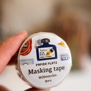 Op de afbeelding: Een rol Papier Platz masking tape met een witte achtergrond. De tape bevat illustraties en tekst, waaronder "hello? BLUE BLACK SMALL THINGS!" en "Masking tape W20mmx5m". De tape wordt in een hand gehouden.
