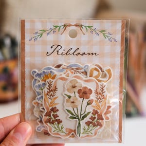 May include: A package of floral stickers with the word "Ribloom" printed on the packaging. The stickers feature watercolor illustrations of flowers in shades of brown, orange, and blue. The background is a light brown gingham pattern.