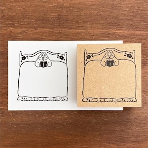 Può includere: Illustrazione in bianco e nero di una persona che legge a letto, con testiera floreale e gonna del letto a balze. L'immagine è su una carta bianca e un'immagine simile su un blocco marrone.