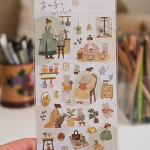 Può includere: Un foglio di adesivi con illustrazioni ad acquerello di persone e conigli in ambientazioni domestiche ed esterne. Le immagini raffigurano cucinare, leggere, giardinaggio e picnic. È presente anche del testo in giapponese e inglese.
