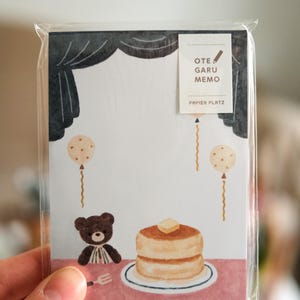 Peut inclure: Bloc-notes sous emballage plastique transparent. Il présente un ours en peluche, des crêpes au beurre et des ballons. Le texte « OTE GARU MEMO » et « PAPIER PLATZ » est également visible. Thème général : mignon et ludique.