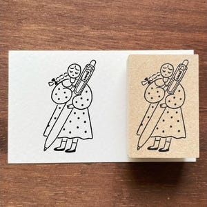 Può includere: Illustrazione in bianco e nero di una persona che tiene una penna, con un vestito a pois e un'espressione triste. Il disegno è su una carta bianca e un timbro di legno.
