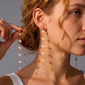 Puede incluir: Elegantes pendientes dorados con perlas colgantes. Los pendientes presentan un pequeño perno dorado en la parte superior, con una delicada cadena que sostiene múltiples perlas. Las perlas son blancas y varían en tamaño, creando un look sofisticado.