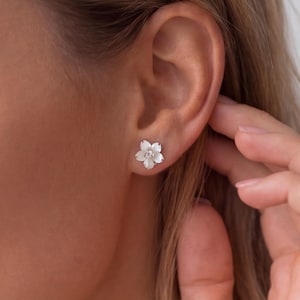 May include: A close-up of a white flower-shaped stud earring with a small, clear gem in the center. The earring is worn on an ear. The earring has five petals and a delicate design.