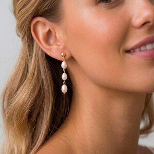 May include: Gold-toned drop earrings featuring three white, teardrop-shaped pearls. The earrings have a small, round gold stud at the top. The pearls are suspended from the stud in a vertical line, creating an elegant and classic look. The earrings are shown on a person's ear.