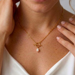 Op de afbeelding: Een goudkleurige ketting met een tuimelsluiting en een druppelvormige parelhanger. De ketting wordt om de hals gedragen en de sluiting is gecentreerd. De ketting is delicaat en de parel is zacht crèmewit.